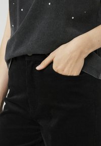 Pantalon en corduroy noir avec une légère texture, associé à un haut sombre avec des éléments réfléchissants. Main reposant dans la poche du pantalon.