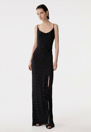 Donna in piedi che indossa un maxi vestito nero senza maniche a pois con uno spacco alto sulla coscia e tacchi neri aperti, su uno sfondo bianco semplice.