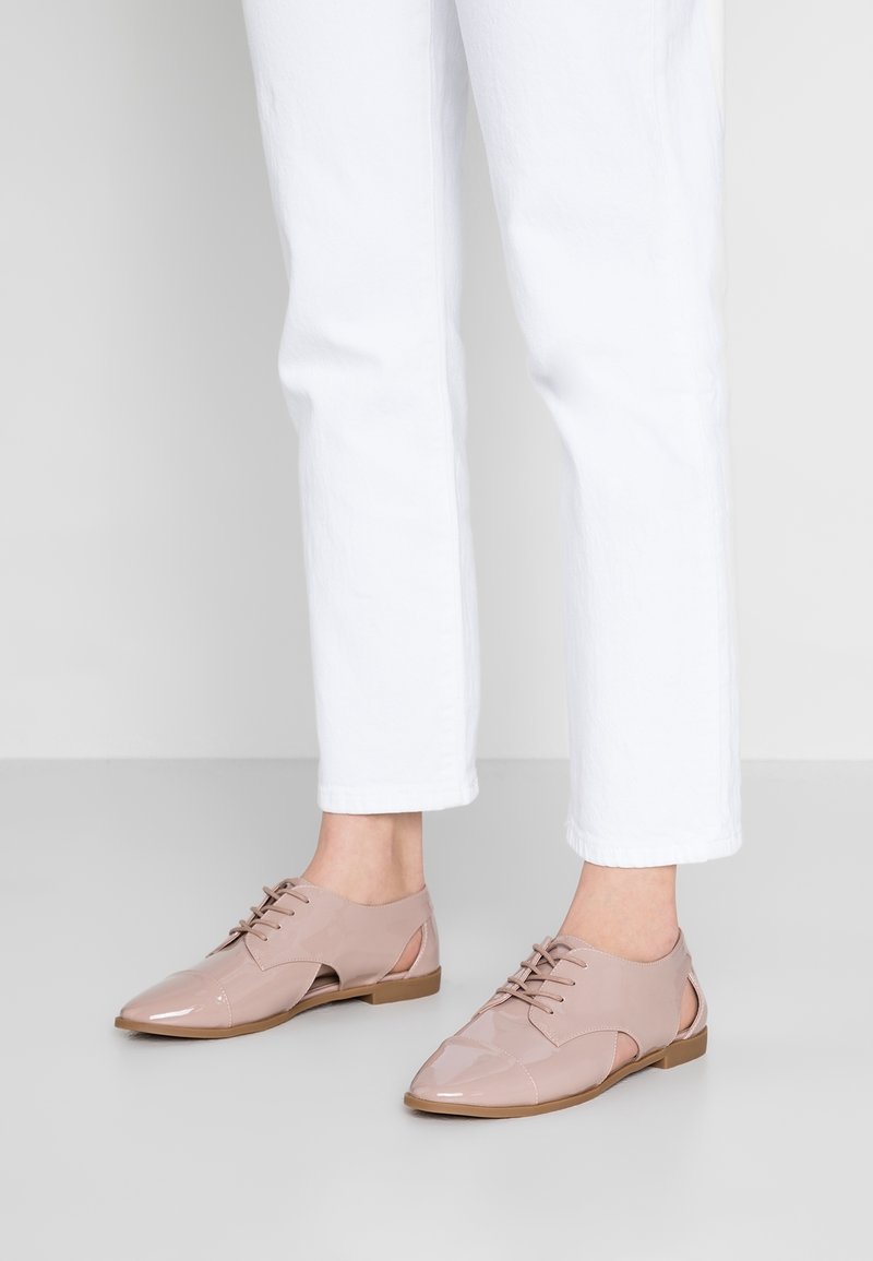 Chaussures à lacets en cuir verni rose clair avec détails découpés, bout arrondi et semelle plate, assorties à un pantalon blanc longueur cheville.