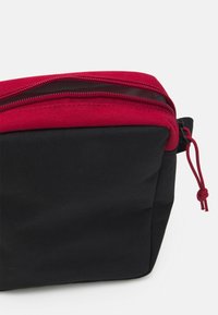 Borsa in tessuto nero con una zip rossa nella parte superiore. Presenta una superficie texturizzata e un dettaglio a cordoncino sul lato, progettata per la conservazione o i viaggi.