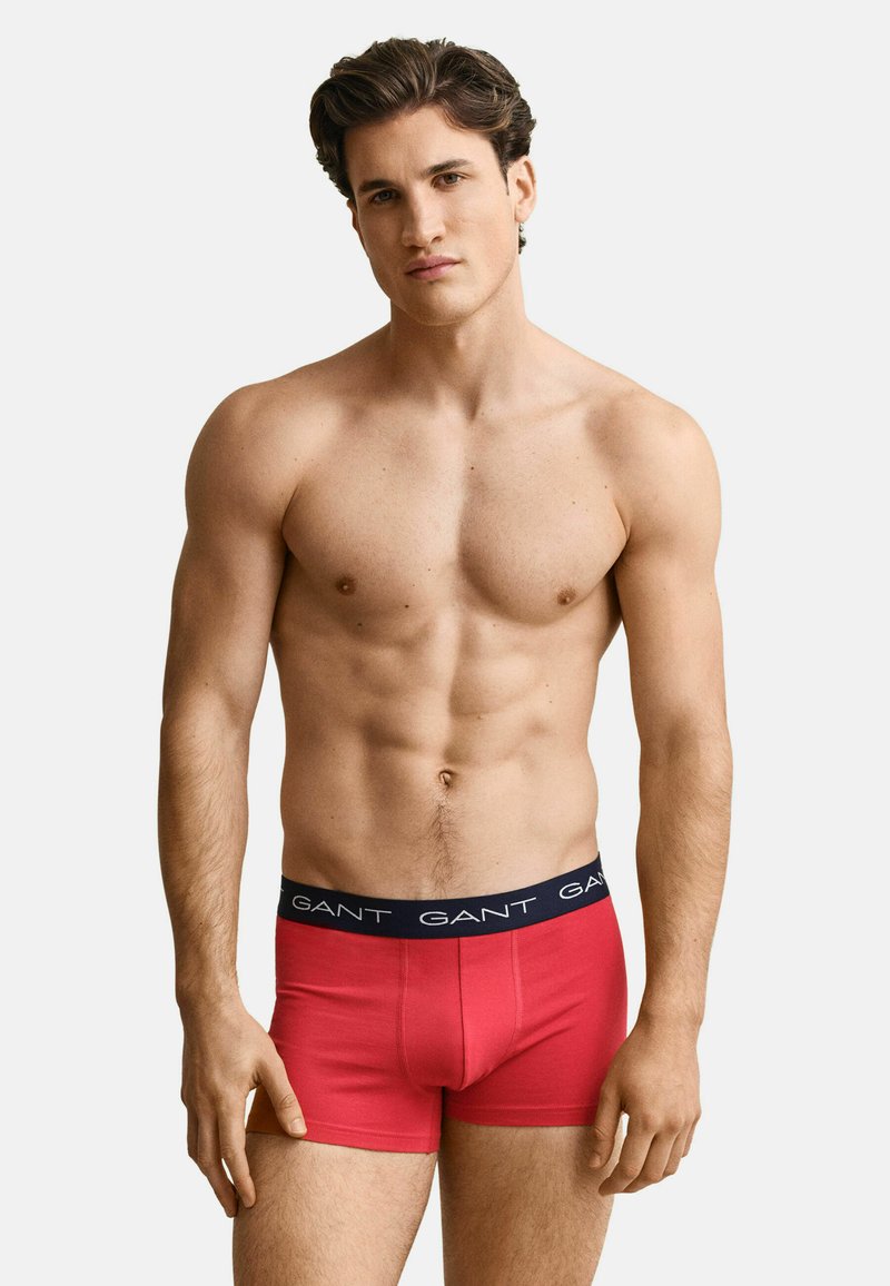 Homme torse nu aux cheveux courts et foncés portant un boxer rouge GANT, debout devant un fond clair uni.