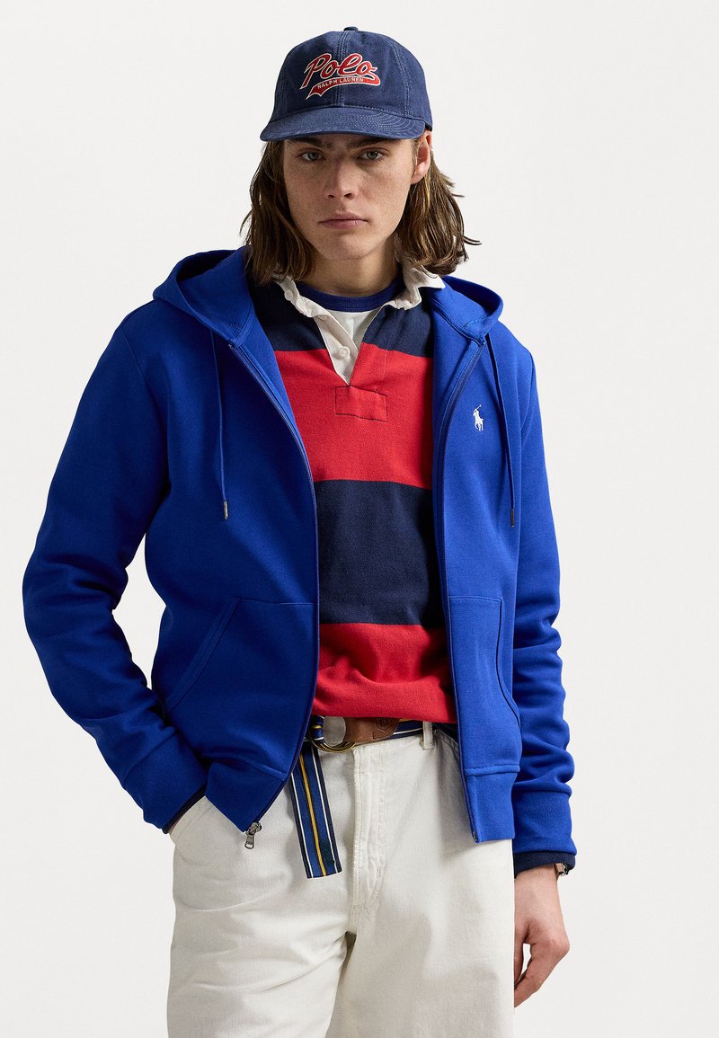 Jovem com cabelo até aos ombros, usando uma basebol azul, um hoodie azul de fecho, uma camisa de rugby listrada em vermelho e azul-marinho, e calças brancas.