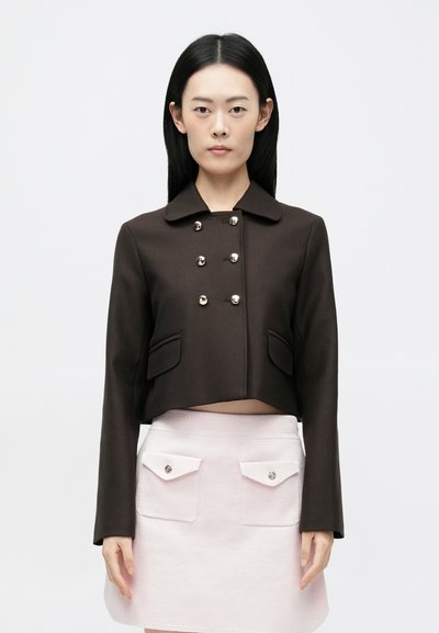 Bruine cropped jas met een kraag, voorzien van zilveren knoopdetails en twee zijzakken, gecombineerd met een lichtroze rok met zakaccenten.
