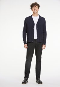 Marinblå stickad kofta med knappknäppning, två sidofickor, bärs över en vit T-shirt, ihop med svarta byxor och svarta stövlar.