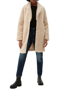 Manteau oversize en fausse fourrure beige clair avec un col châle, deux boutons, poches latérales et une texture douce, associé à un jean bleu foncé et des bottes noires.