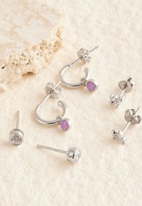 Pendientes de plata que incluyen estilos de aro con una piedra púrpura y múltiples diseños de tachuelas, presentando formas redondas y florales, todos exhibidos sobre un fondo suave.