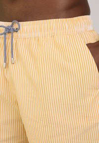 Shorts rayés en tissu léger, avec des rayures jaunes et blanches, dotés d'une taille élastique avec cordon de serrage et d'un détail de poche sur le côté.