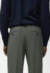 Persona con una camiseta de manga larga azul marino oscuro, metida dentro de unos pantalones de cintura alta de color verde oliva con bolsillos traseros abotonados, vista desde atrás.