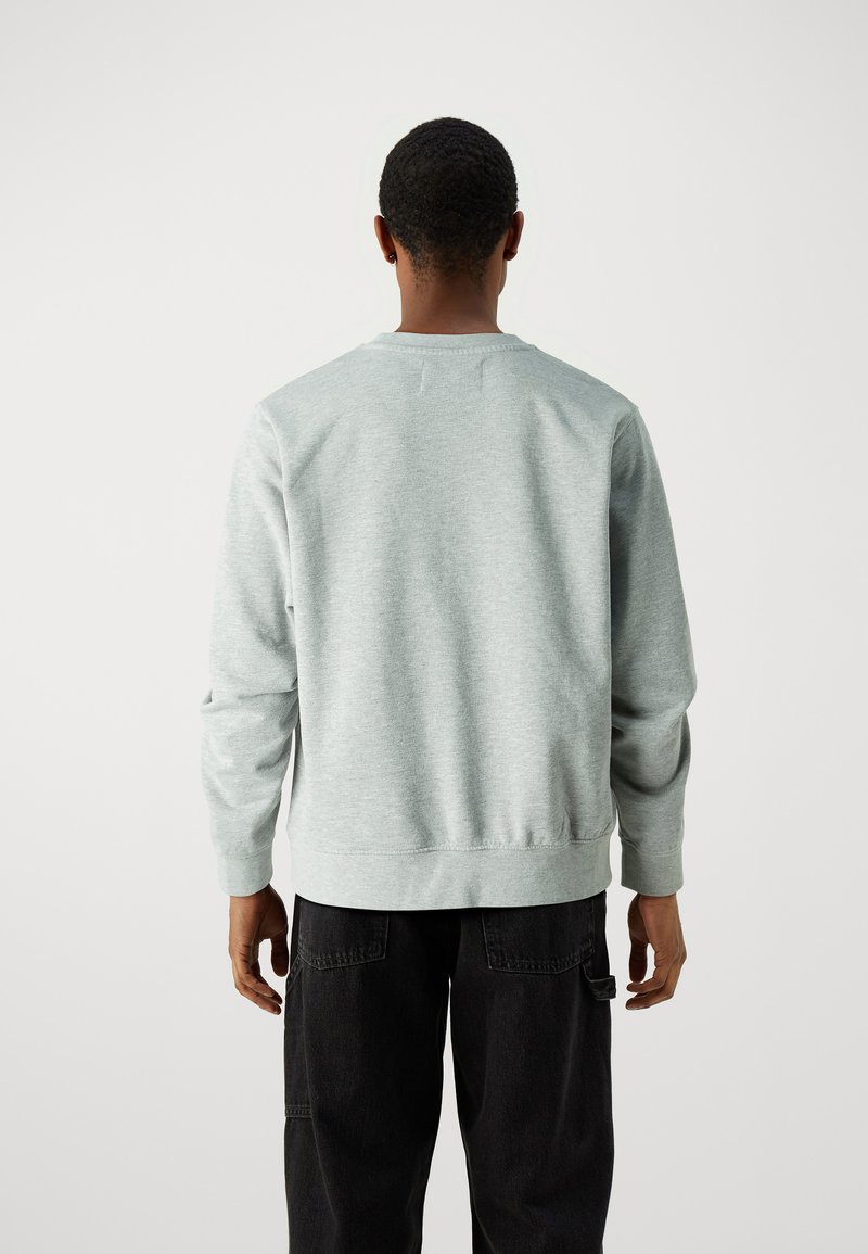 Sudadera gris claro con escote redondo y puños acanalados. Textura suave, corte clásico, vista desde la parte posterior con detalles mínimos. Pantalones negros visibles.