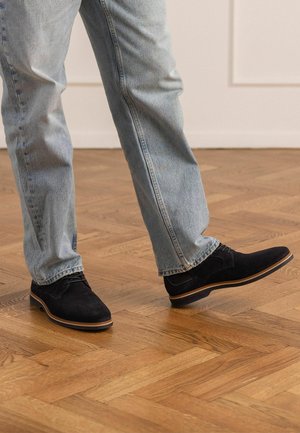Persoon draagt lichtblauwe spijkerbroek en zwarte suède veterschoenen, staand op een houten parketvloer.