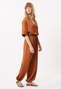 Roestkleurige jumpsuit met korte mouwen, V-hals, elastische taille en taps toelopende pijpen. Model draagt platte sandalen en een gepatternde hoofddoek.