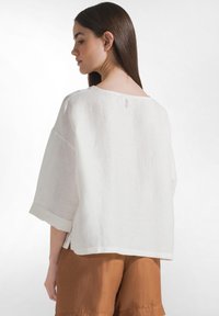 Kvinde med langt, mørkt hår iført løs, hvid bluse med opkrullede ærmer og brune shorts, vist bagfra mod en ensfarvet baggrund.