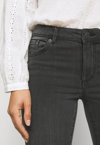 Dunkelgraue Jeans aus Denim mit enganliegendem Schnitt, einer Vorderseite mit Tasche und einem Metallknopfverschluss. Dazu eine weiße, strukturierte Bluse.