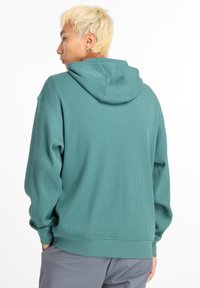 Sudadera con capucha color azul verdoso con canalé, capucha con cordón, ajuste holgado, mangas largas y superficie texturizada, combinada con pantalones grises.