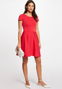 Rotes, tailliertes Kleid mit kurzen Ärmeln und ausgestelltrock, aus strukturiertem Stoff. Kombiniert mit einer weißen Clutch und spitzen Ballerinas.