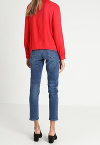 Blusa rossa con maniche lunghe e schiena a occhietto; abbinata a jeans skinny blu e mules nere con tacco. Tessuto trasparente, vestibilità casual.