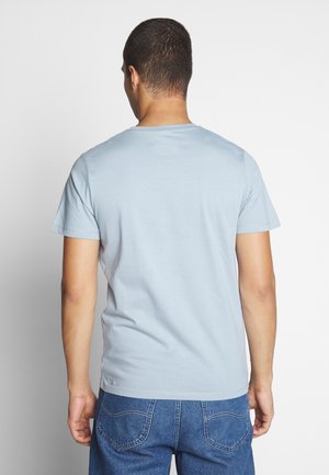 Homme portant un t-shirt à manches courtes bleu clair et un jean bleu, se tenant de dos face à la caméra sur un fond blanc uni.