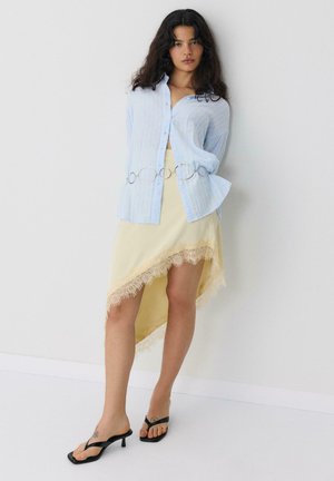 Žena v svetlomodrej košeli na gombíky so strieborným opaskom, asymetrickej bledozelenej sukni s čipkovaným lemom a čiernych sandáloch na podpätku, stojaca.