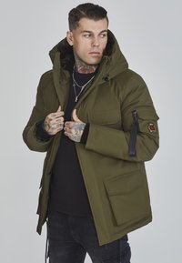 Oliven grøn parka med en foring af pels i huen, frontlynlås og lommer; har et logo-patch på ærmet og strukturerede detaljer.