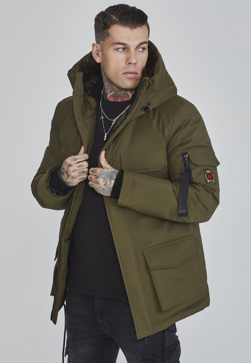 Oliven grøn parka med en foring af pels i huen, frontlynlås og lommer; har et logo-patch på ærmet og strukturerede detaljer.