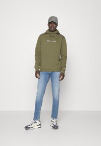 Sudadera verde oliva con un bolsillo canguro, que presenta un logo blanco en el pecho, combinada con jeans de color azul claro y zapatillas multicolores.