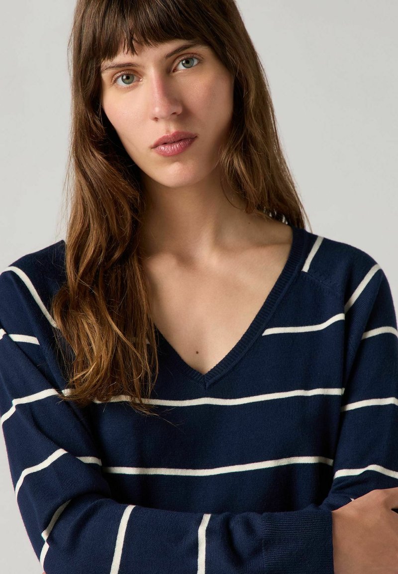 Pull en V bleu marine en tissu avec des rayures horizontales blanches. Il dispose de manches longues et d'une coupe décontractée. Texture douce avec des poignets côtelés.