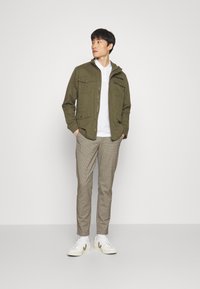 Oliven grøn jakke med flere lommer, hvid polo shirt, beige ternede bukser og hvide sneakers. Glat stof med en afslappet pasform.