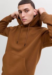 Ung mand justerer hætten på en brun sweatshirt med frontlomme mod en ensfarvet lys baggrund.