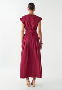 Dlhé burgundské šaty s krátkymi rukávmi, priliehavý pás zvýraznený opaskom a skladaným chrbtom. Hladká látka a dizajn po členky.