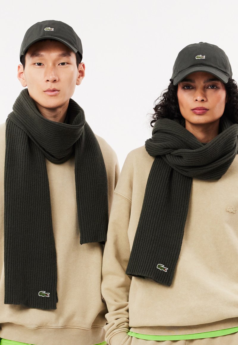 Sciarpa a coste verde scuro indossata da due modelli, abbinata a felpe beige con un piccolo logo e un accento verde. Cappelli scuri completano il look.