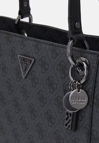Sac à main noir Guess avec motif de logo embossé, emblème triangulaire en métal Guess USA, et breloques métalliques rondes et rectangulaires Guess attachées.