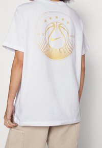 Bílý bavlněný tričko s krátkým rukávem, na zadní straně je zlatá grafika basketbalového míče a pěti hvězd, označeno "1972-2023."