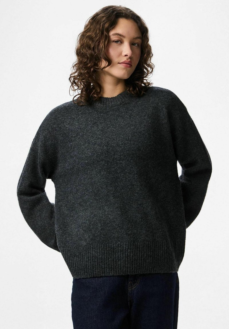 Ung person med lockigt hår som bär en mörkgrå stickad tröja och mörka byxor, stående med händerna bakom ryggen mot en vit bakgrund.