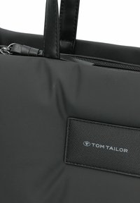Zwarte stoffen tote bag met een gladde textuur, voorzien van leren handvatten en een textuurpatch met het "TOM TAILOR" logo. Ritsafsluiting aan de bovenkant.