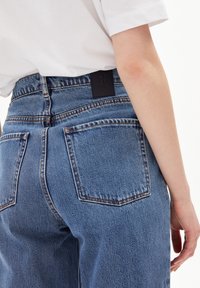 Jeans de mezclilla azul con un corte relajado, diseño de cinco bolsillos y una etiqueta de marca de cuero negro en la parte posterior. Costuras visibles que destacan en todo el diseño.