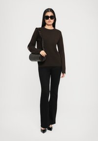 Woman wearing black sunglasses, dark brown sweater, black flared pants, black pointed heels, carrying a black cylindrical handbag on shoulder.