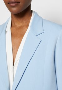 Blazer azul claro con hombros estructurados, solapas de muesca y una textura suave, acompañado de una camisa blanca con cuello por debajo.