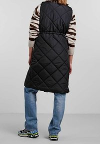 Svart kviltad lång väst med åtdragen midja, bärs över en randig tröja, kombinerad med blå jeans och färgglada träningsskor.