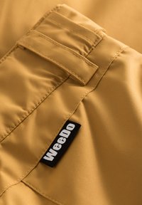 Tan tyg med en slät yta, som har en klaffficka och en svart "WeeDo"-etikett. Sömnadsdetaljerna förstärker strukturen och hållbarheten.