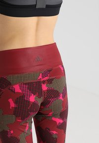 Leggings de tiro alto en color marrón burdeos con un patrón de camuflaje geométrico en rosa y marrón. Presentan una textura suave y el logo de Adidas en la cintura.