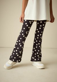 Pantalon noir évasé à grands pois blancs, en tissu côtelé, associé à des baskets de couleur claire avec une semelle texturée.