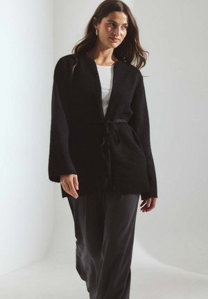 Schwarze, strukturierte Strickjacke mit Wickeldesign, weiten Ärmeln und einer gebundenen Taille, kombiniert mit grauen Hosen. Bietet einen lässigen, lockeren Schnitt.