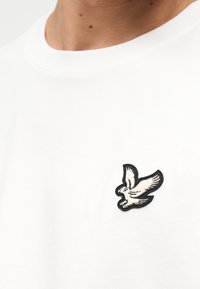 Gros plan sur une personne portant une chemise blanche avec un petit oiseau blanc brodé et un contour noir sur la poitrine.