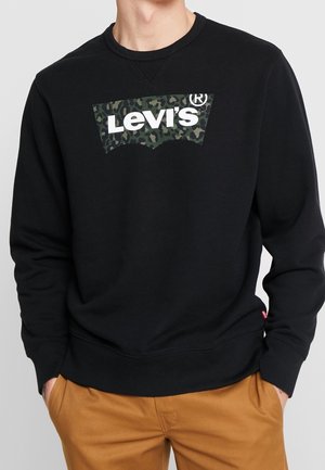 Person trägt schwarzen Levi's Sweatshirt mit grünem Camouflage-Logo und hellbraunen Hosen, Hände in den Taschen, steht vor einfachem Hintergrund.
