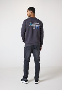 Mørkegrå sweatshirt med broderet tekst og blå sommerfugledesign på ryggen, parret med mørke jeans og sort-hvide sneakers.