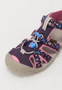 Lila und rosa Sandale mit einem Blumenmuster, verstellbaren Riemen, einem Schnellverschluss-System und einer beigen Gummisohle. Waschbares Material.