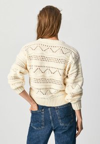 Kvinna iklädd en krämfärgad stickad tröja med öppenmönster och blå denimjeans, står med en hand i bakfickan.