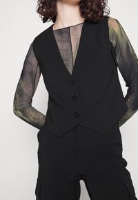 Schwarze taillierte Weste mit V-Ausschnitt, drei Knöpfen und seitlichen Taschen, kombiniert mit einer langärmeligen, durchsichtigen Bluse mit Muster.