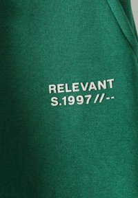 Zaļš audums ar gludu tekstūru, kam priekšā ir balts izšuvums ar tekstu: "RELEVANT S.1997 //--".