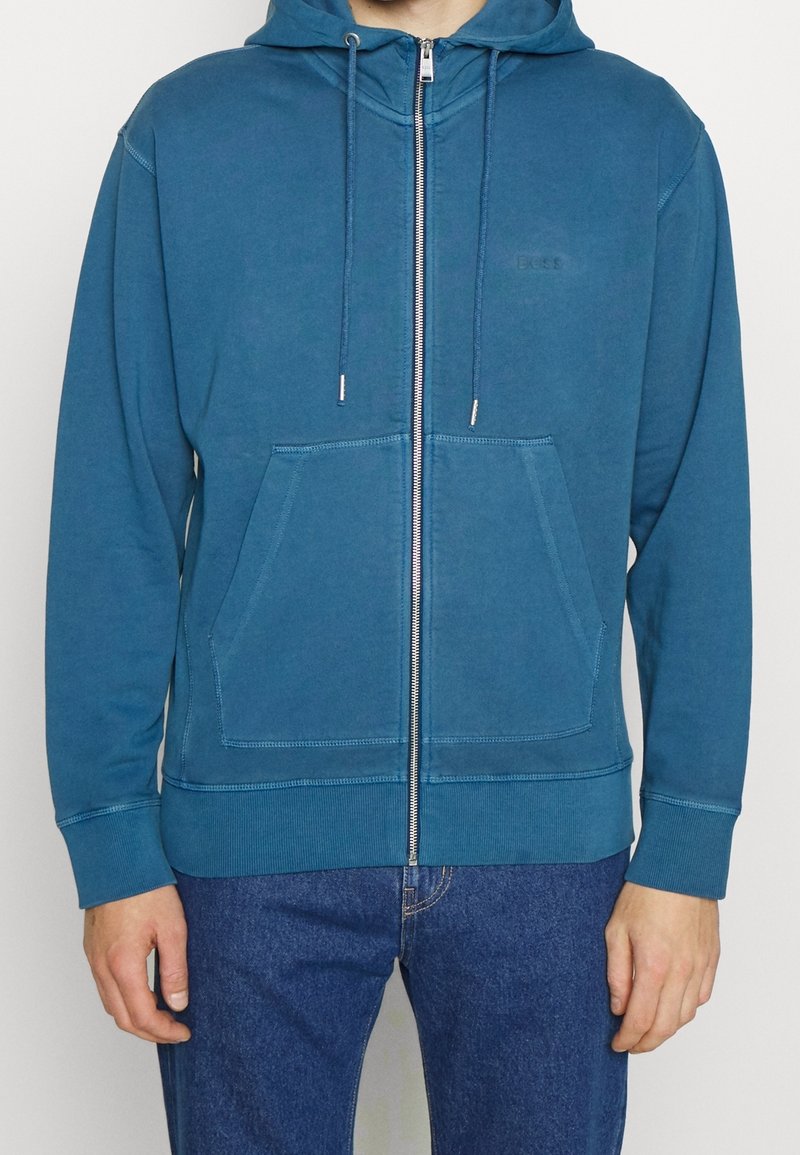 Homme portant un sweat à capuche zippé bleu sarcelle avec poches avant et cordons, associé à un jean bleu foncé, montré des épaules aux hanches.